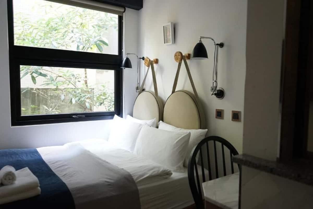 A neatly made bed with white linens and a blue accent, a window with a view of greenery, and two wall-mounted lamps.