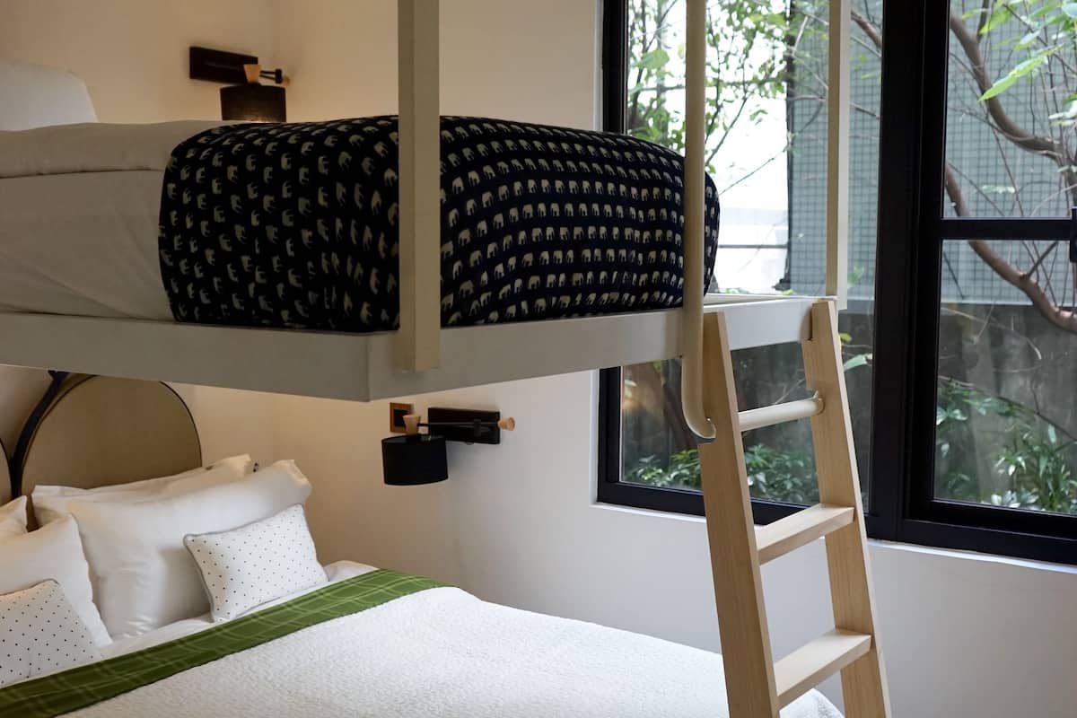 A bunk bed with a ladder, a bed with white bedding, and a window with a view of trees.