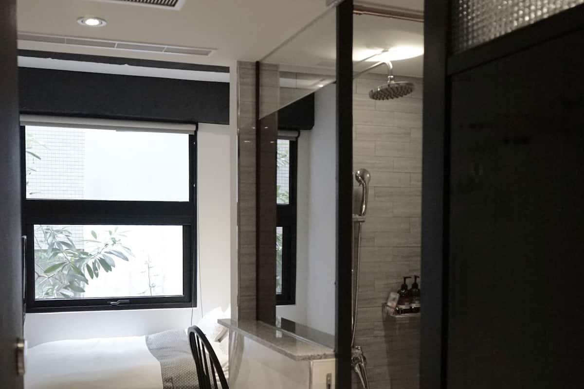 A modern hotel room with a bed, a chair, a shower area, and a large window.