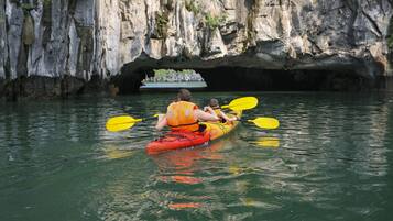 A kayak with two people paddling on calm water, a rocky cliff in the background, and a boat visible through a cave opening.