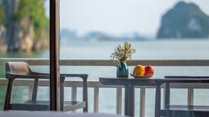 A table with a vase of flowers and a plate of fruit, overlooking a body of water with a distant island.
