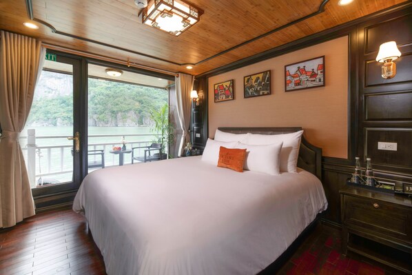 A hotel room with a large bed, a wooden floor, a balcony with a view of greenery, and artwork on the wall.