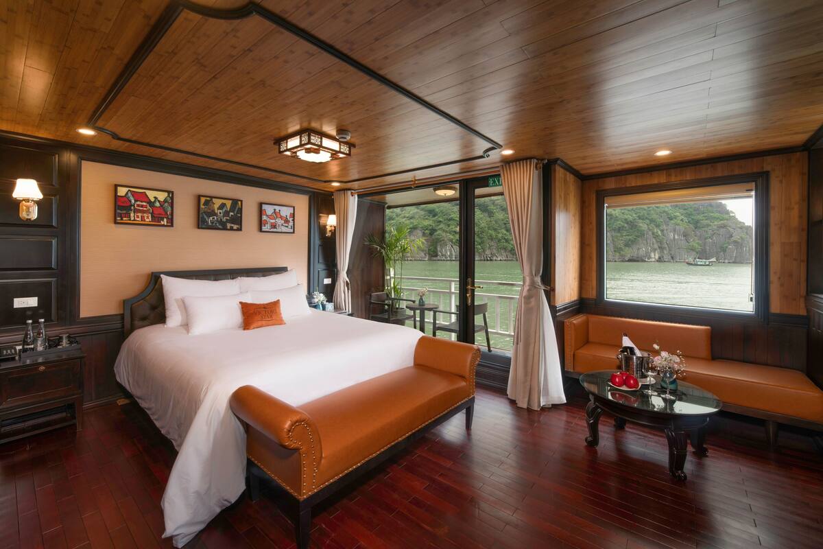 A spacious room with a large bed, a seating area by a window, and a balcony with a view of the water.