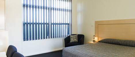 A hotel room with a bed, a chair, a nightstand, and a window with blinds.