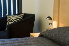 A hotel room with a dark sofa, a zebra-patterned pillow, a bedside table with a lamp, and a neatly made bed. - Cherry Blossom Motel (Young)