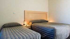 Quarto de hotel com duas camas de solteiro, cabeceira de madeira, mesas de cabeceira com abajures e uma parede branca lisa.