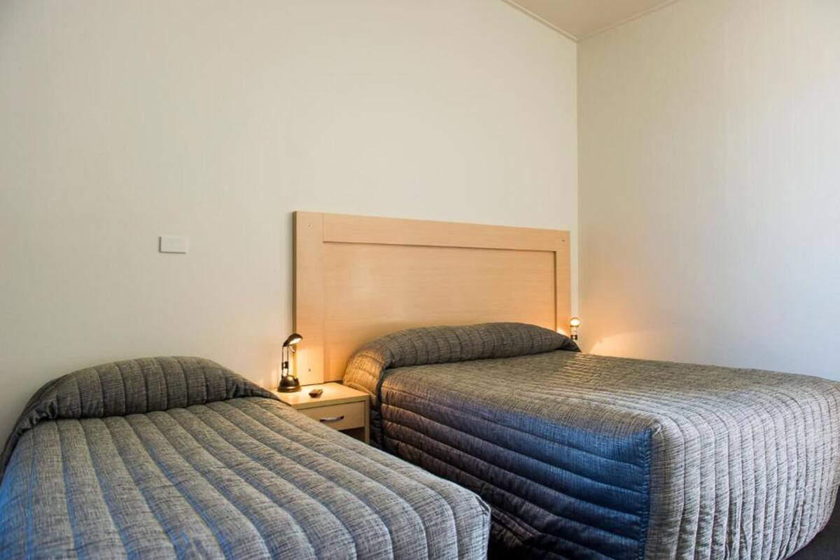 A hotel room with two single beds, a wooden headboard, bedside tables with lamps, and a plain white wall.