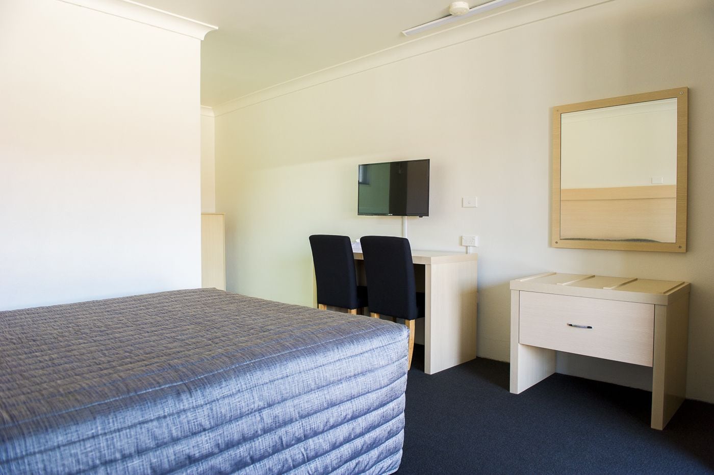 A hotel room with a bed, a desk with two chairs, a television, and a bedside table with a mirror.