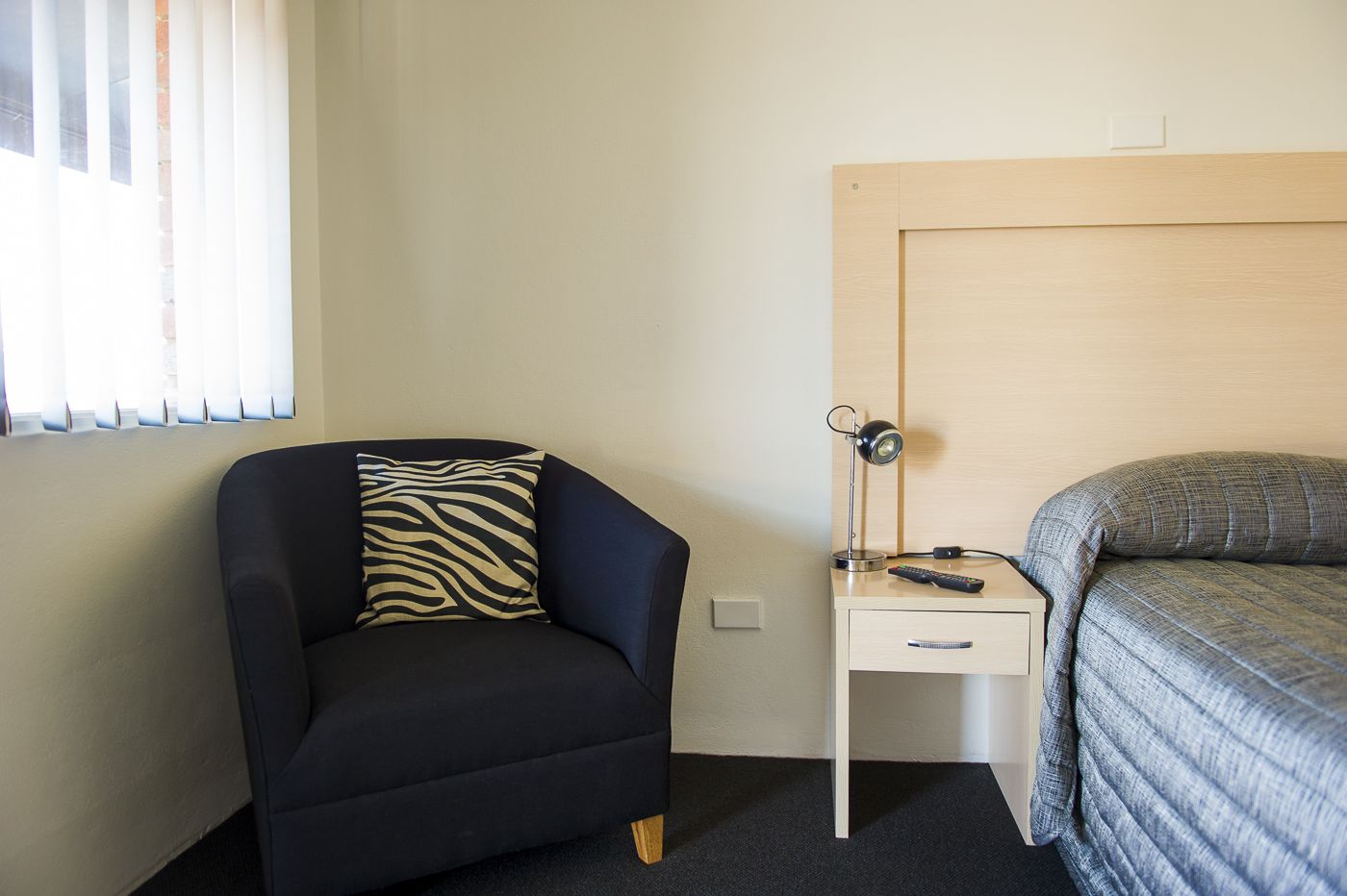 Una habitación de hotel pequeña con una cama individual, una silla con un cojín estampado de cebra, una mesita de noche con una lámpara y mandos a distancia, y una ventana con persianas.