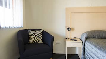 Una habitación de hotel pequeña con una cama individual, una silla con un cojín estampado de cebra, una mesita de noche con una lámpara y mandos a distancia, y una ventana con persianas.