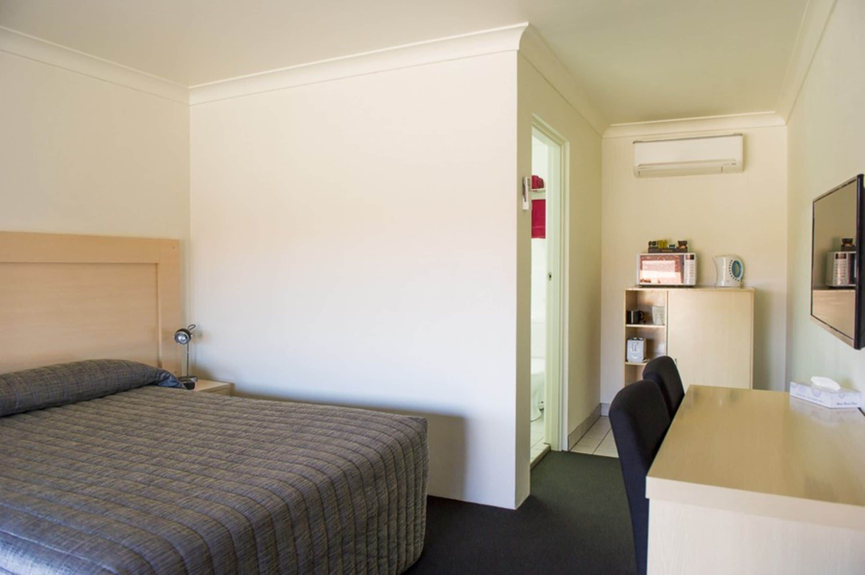 A hotel room with a bed, a desk, a chair, a nightstand, a shelf with toiletries, and a door leading to a bathroom.
