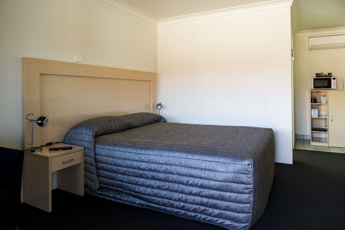 A single bed with a headboard, a bedside table with a lamp, and a small shelf unit with a microwave.