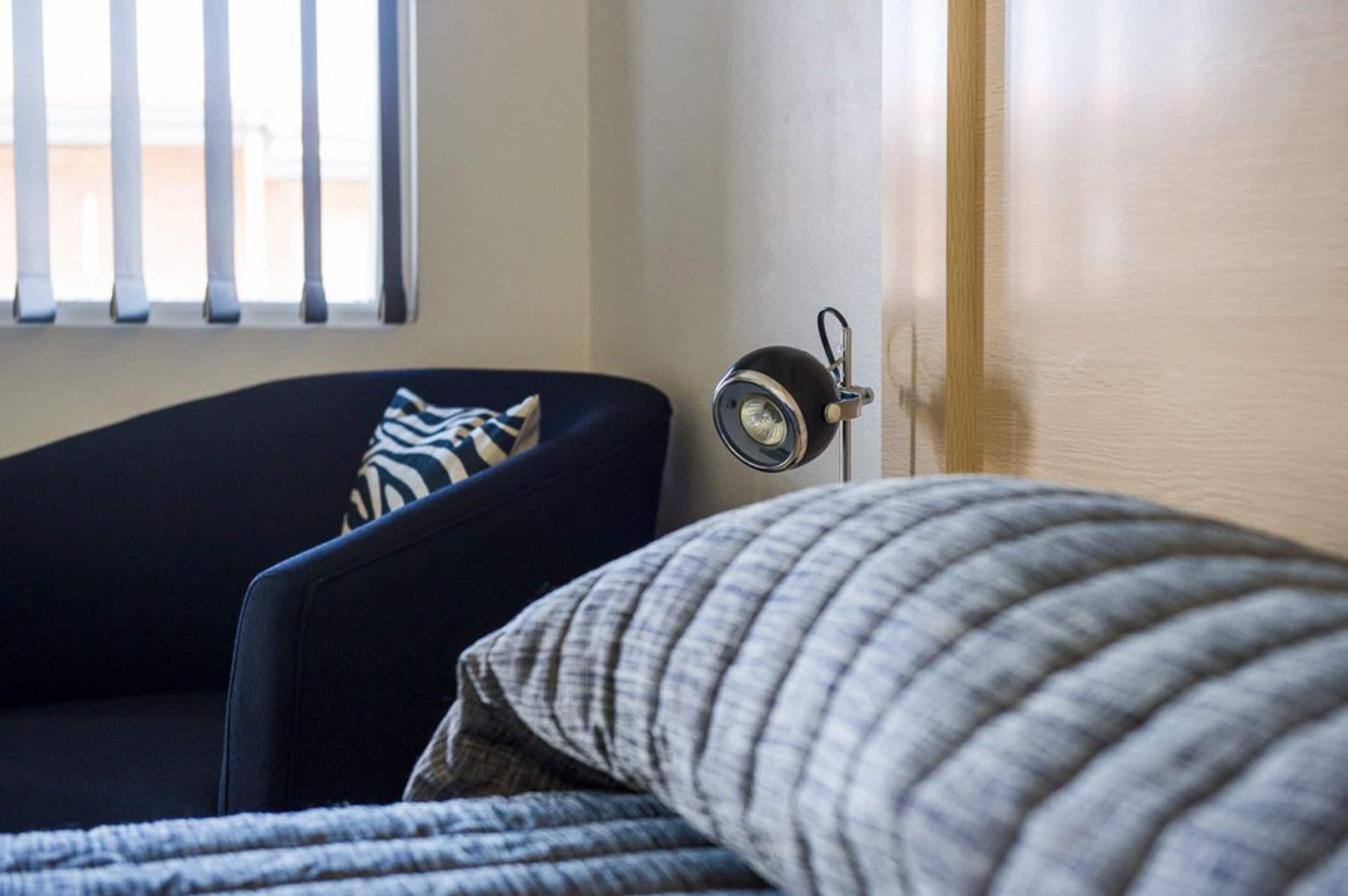 A room with a dark blue armchair, a striped pillow, and a lamp on a bedside table.