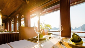 Mesa de comedor servida con platos, cubiertos y un vaso, con vista al mar y montañas de fondo.