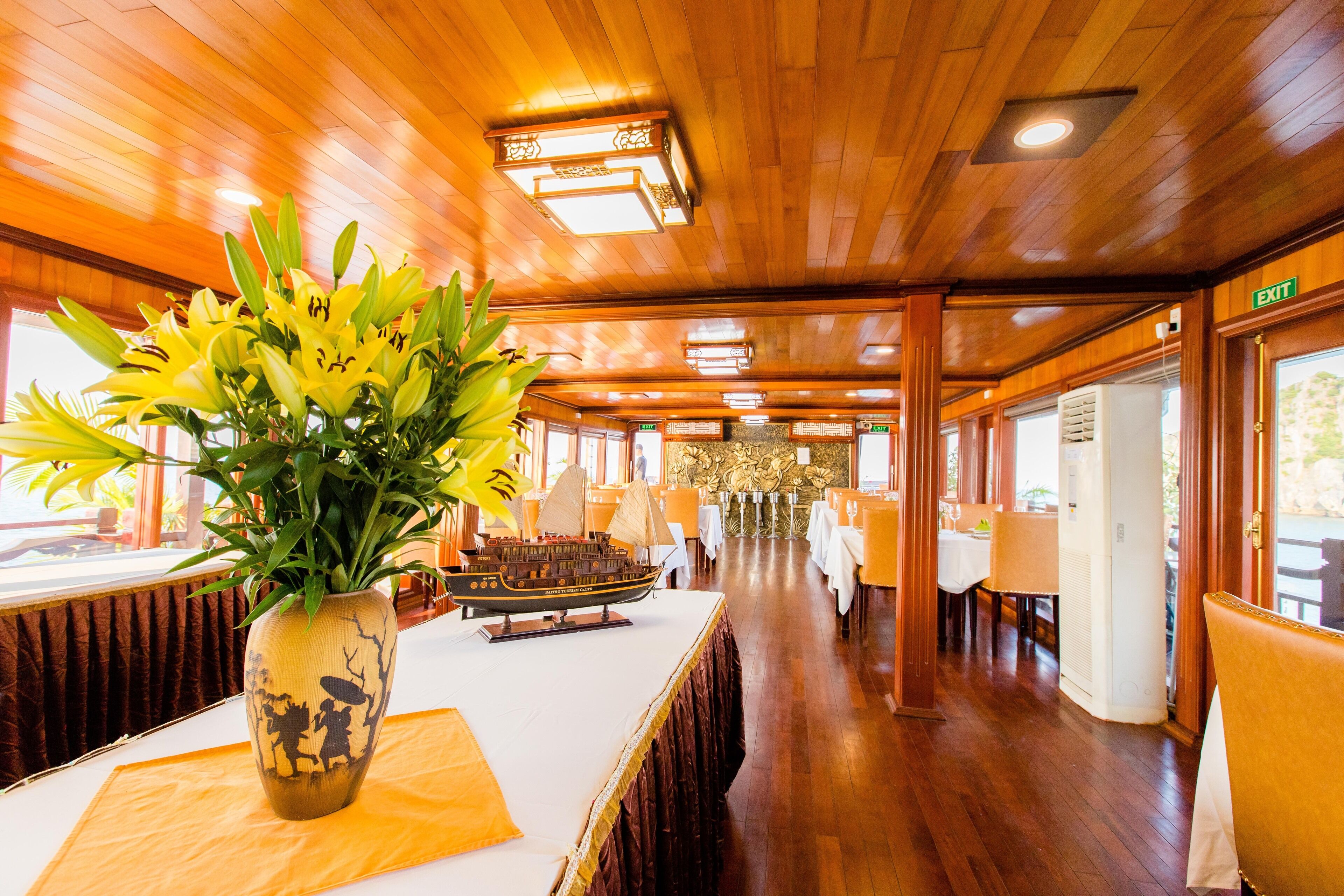 A wooden interior with a long table, a vase with yellow flowers, and a ship model.