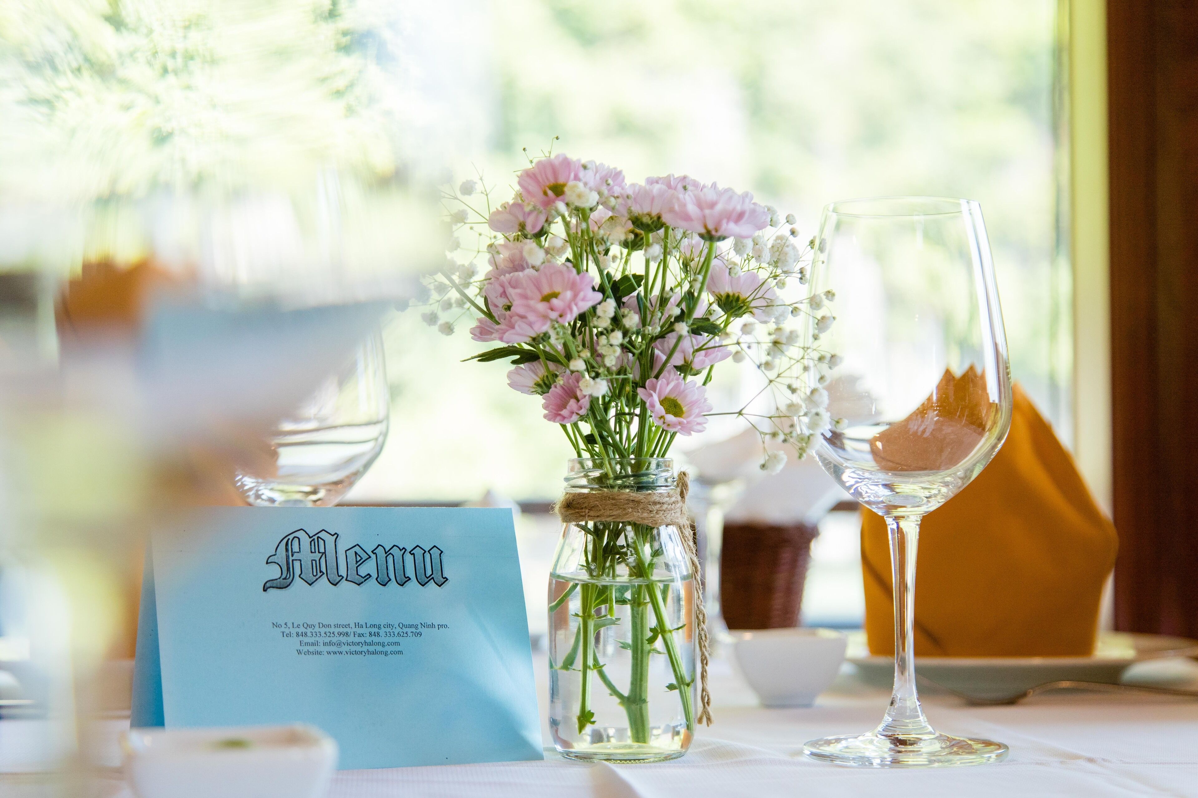 A table with a clear glass jar containing pink flowers, a wine glass, and a menu card.