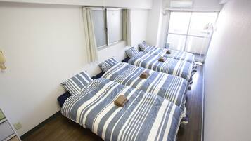 A hotel room with three beds, each with striped bedding and towels. There is a window with curtains, a wooden floor, and a white wall.