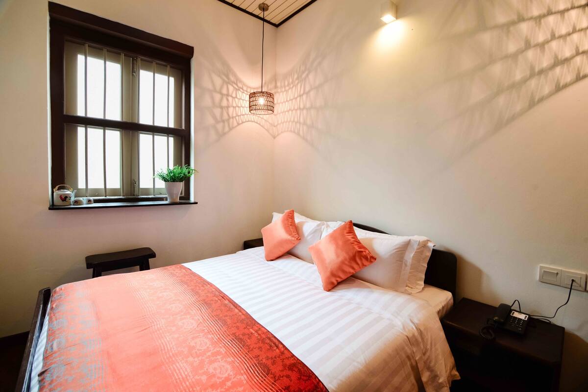 Un lit bien fait, recouvert d’une couvre-lit à motifs, avec deux oreillers, une table de chevet sur laquelle se trouve un téléphone, et une fenêtre avec des stores qui projettent des ombres sur le mur.