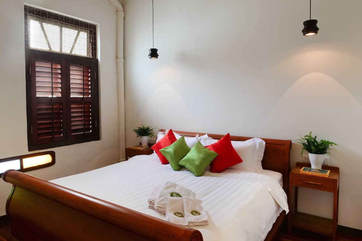 Un lit bien fait, avec du linge de lit blanc et des coussins verts et rouges, une tête de lit en bois et deux tables de chevet ornées de plantes.