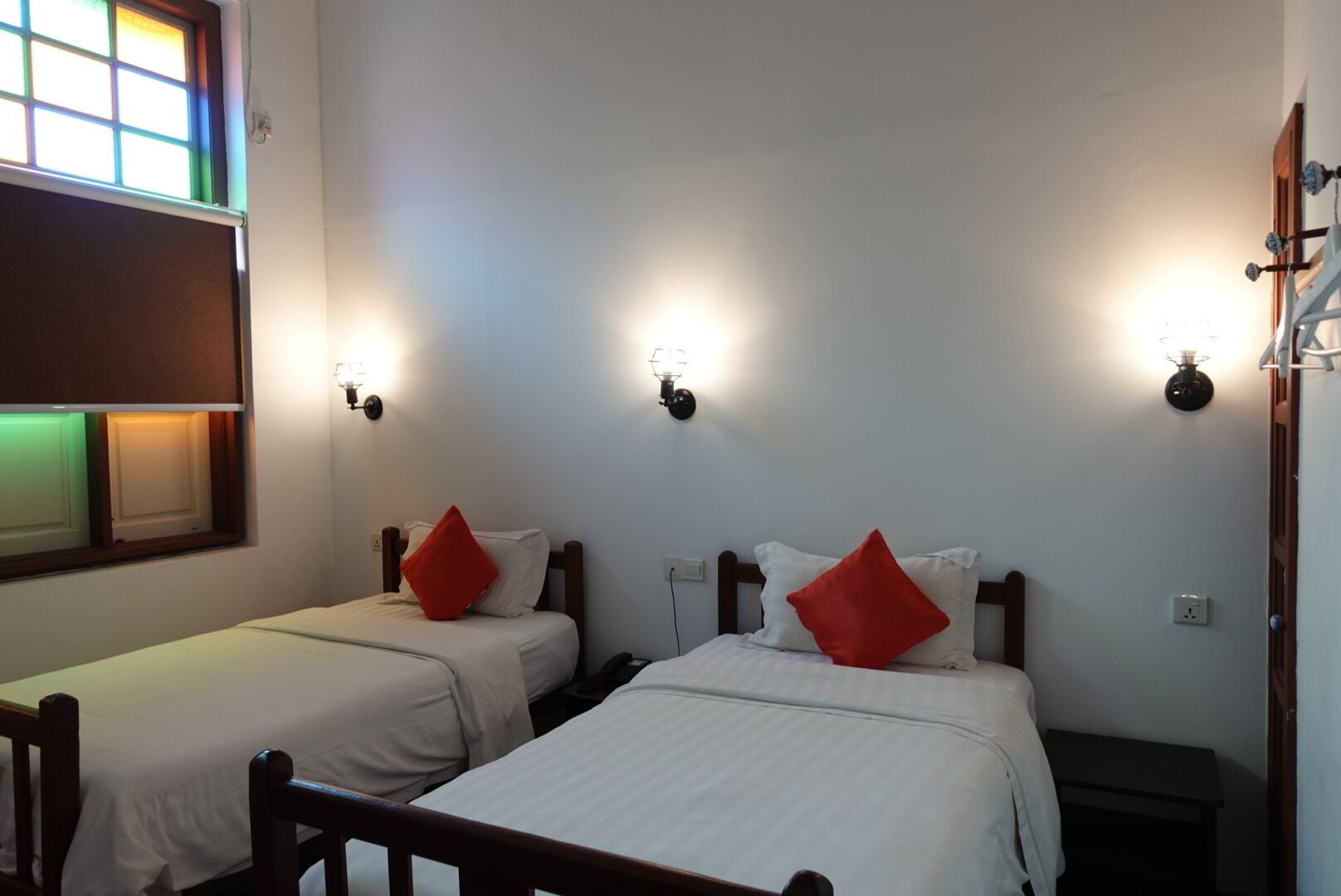 Dos camas individuales con cabeceros de madera, ropa de cama blanca y almohadas rojas. Cada cama tiene una mesita de noche con una lámpara. La habitación tiene paredes blancas y una ventana con vistas al exterior.