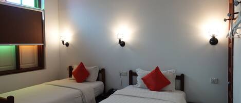 Dos camas individuales con cabeceros de madera, ropa de cama blanca y almohadas rojas. Cada cama tiene una mesita de noche con una lámpara. La habitación tiene paredes blancas y una ventana con vistas al exterior.