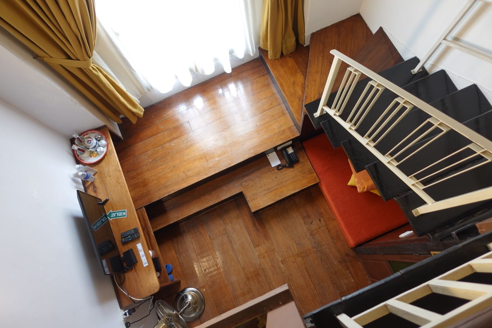 A room with a wooden floor, a bed, a desk with a chair, a small table with a plate and cups, and a staircase with a railing.