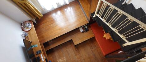 A room with a wooden floor, a bed, a desk with a chair, a small table with a plate and cups, and a staircase with a railing.