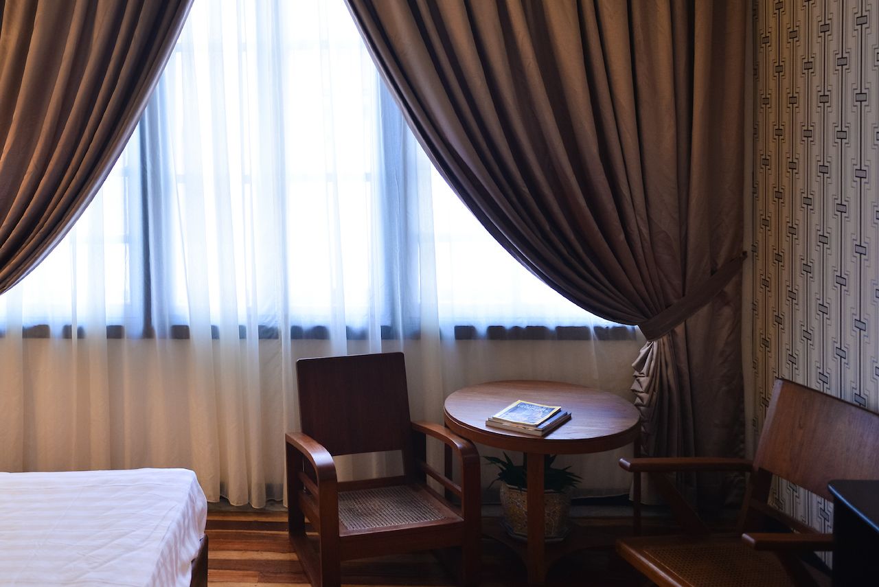 A hotel room with a bed, two wooden chairs, a small round table with a book, and a window with sheer curtains.