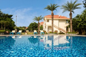 A swimming pool with blue checkered tiles, lounge chairs, palm trees, and a building with a red roof.