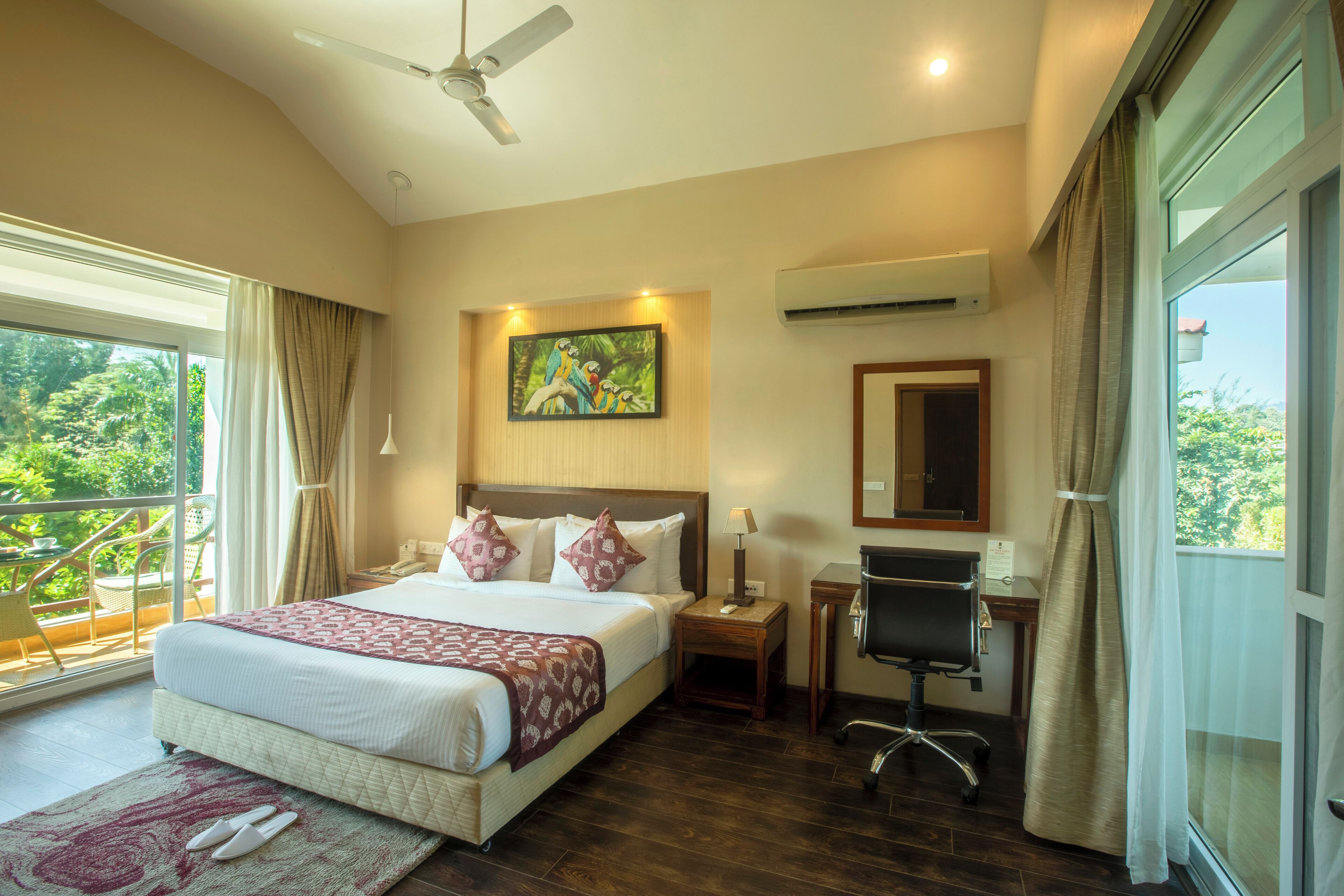 Une chambre spacieuse avec un grand lit, un bureau et une chaise, un ventilateur de plafond et une vue sur la verdure environnante.
