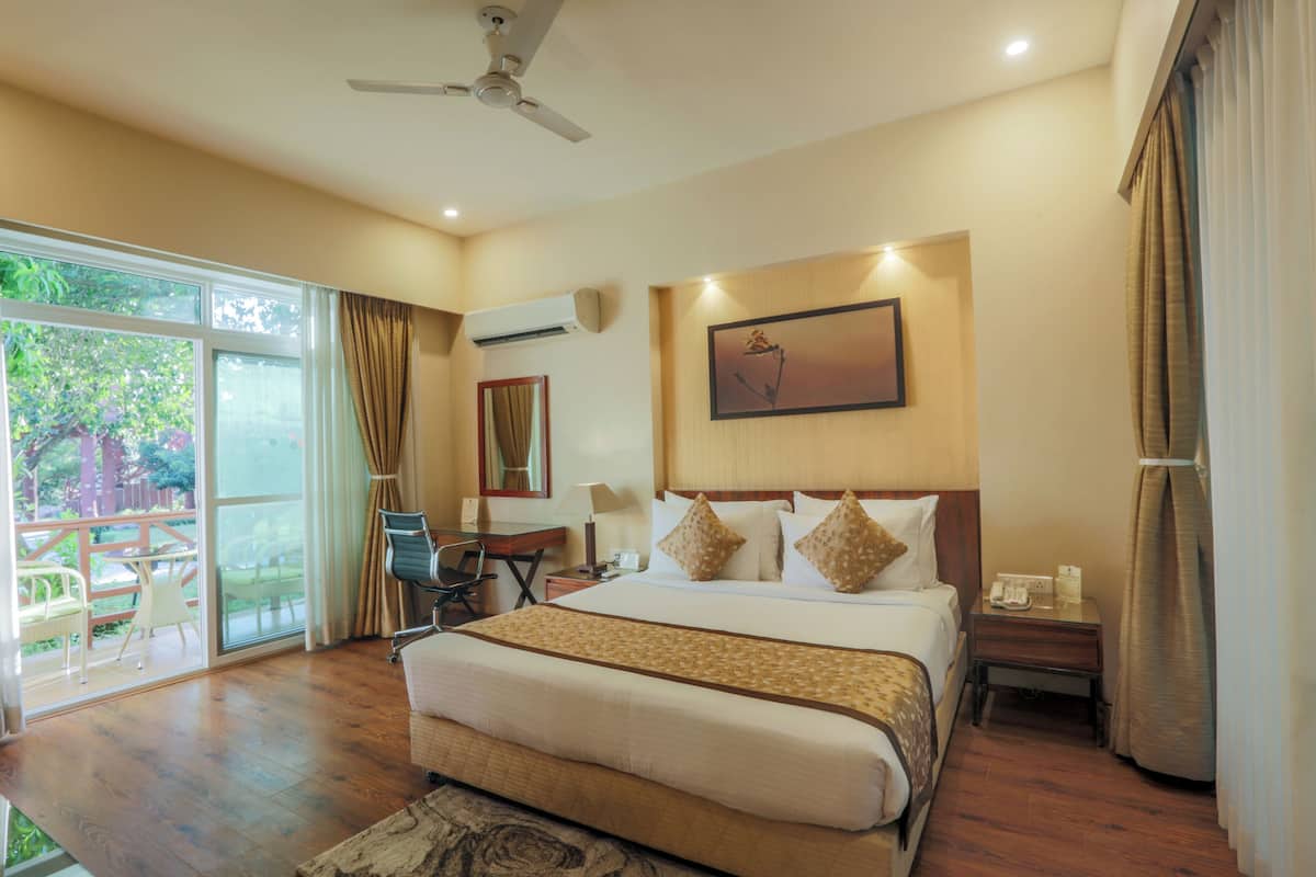 Une chambre d’hôtel avec un grand lit, un bureau et une chaise. Il y a une fenêtre avec des rideaux, un ventilateur de plafond et un tableau au mur.