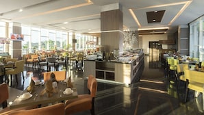 Un amplio comedor de hotel con múltiples mesas y sillas, un mostrador de buffet y ventanas grandes que dejan entrar luz natural.