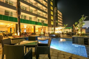 Zona de piscina de hotel con mobiliario exterior, una fuente y un edificio con varios balcones.