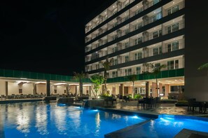 Un hotel de varios pisos con piscina, mobiliario exterior y un vestíbulo bien iluminado.
