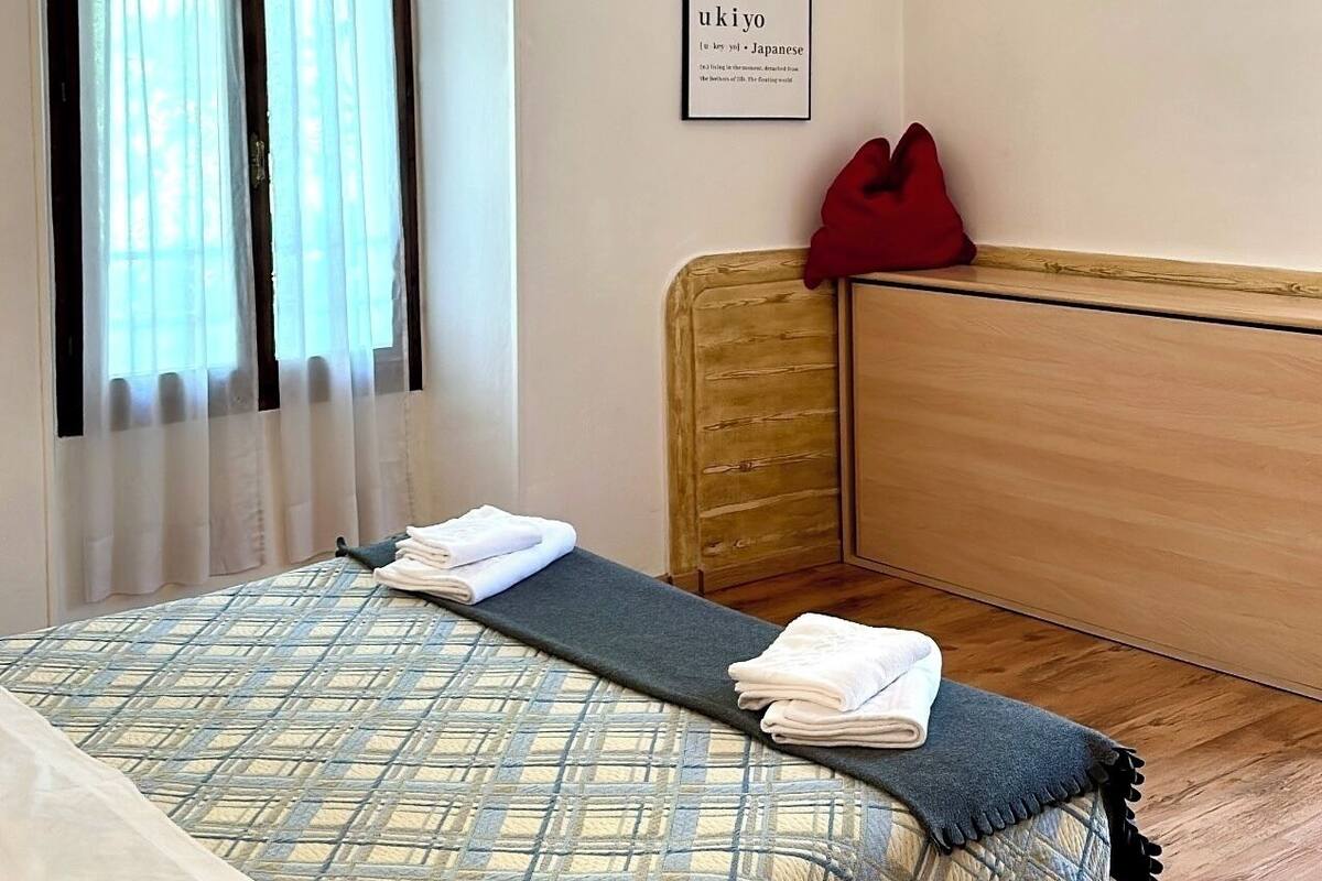 A bedroom with a bed, a window with curtains, a bedside table, a red pillow, and a framed picture on the wall.