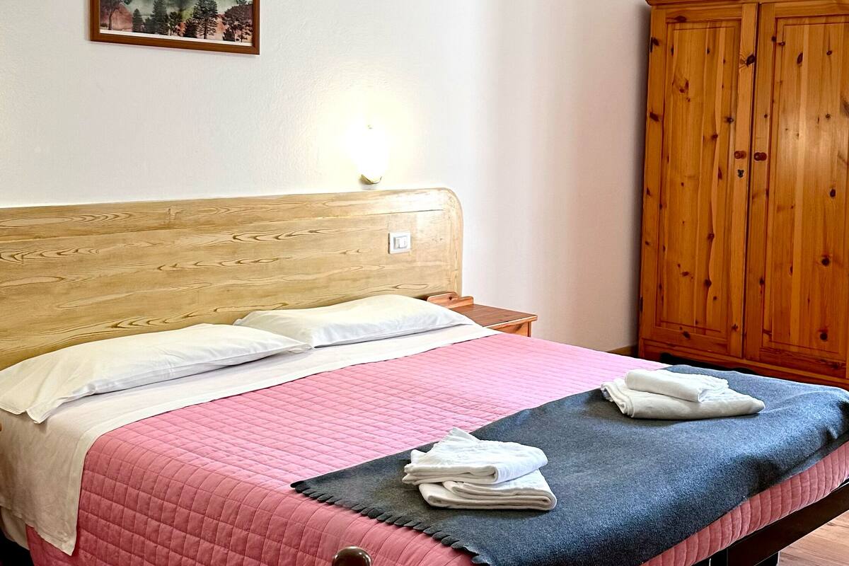 A bedroom with a wooden headboard, a pink quilted bedspread, a wooden table, and a wooden wardrobe.