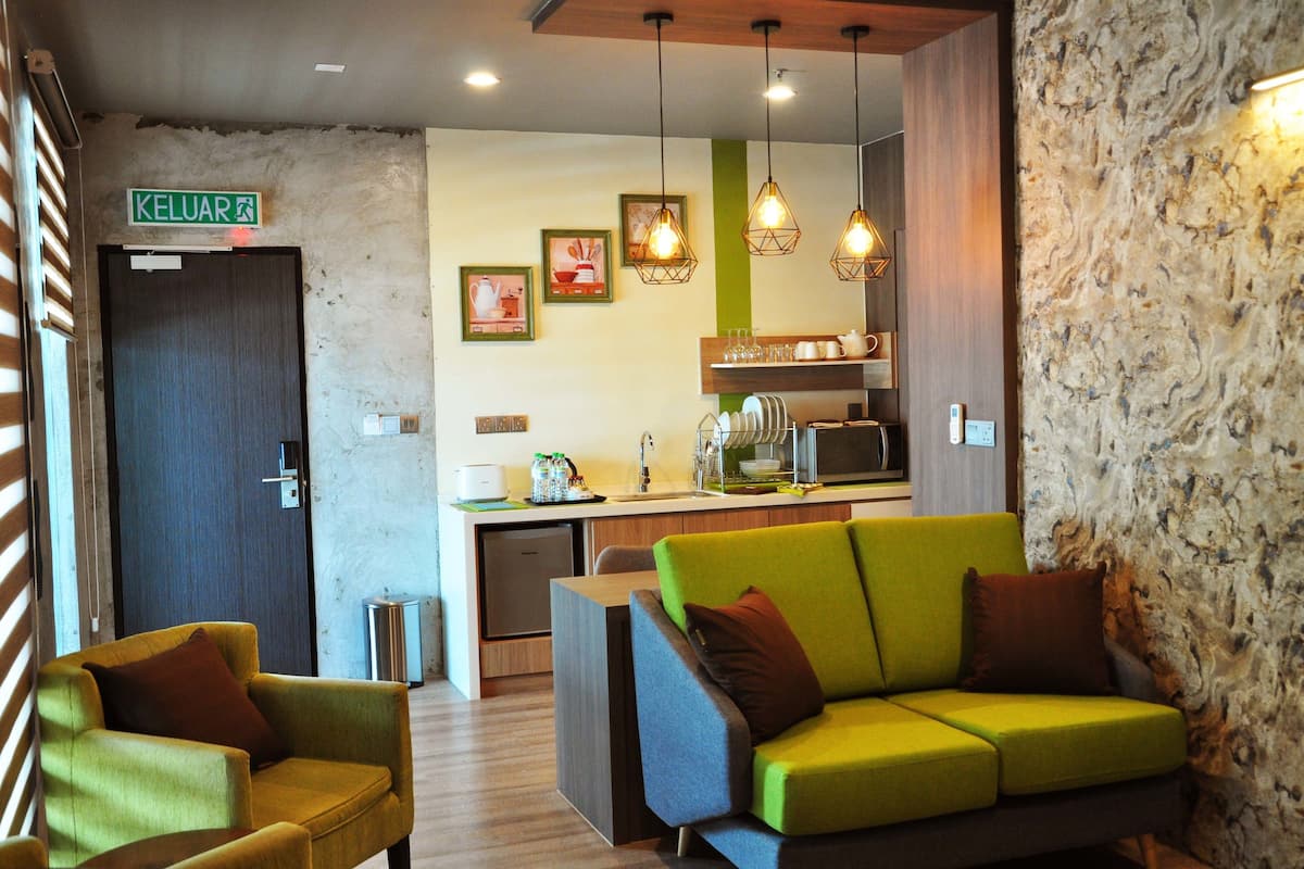 A modern hotel lobby with a green sofa, a wooden-framed door labeled "KELUAR," and a kitchenette area with hanging pendant lights.