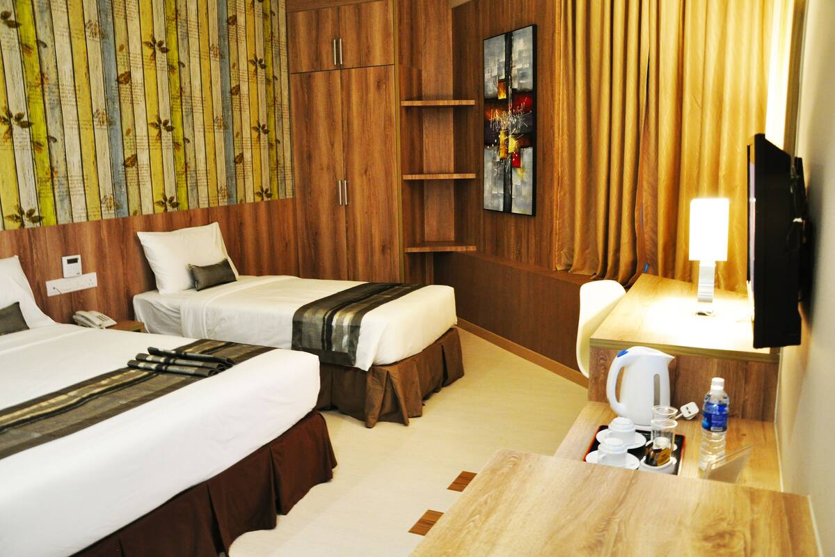 A hotel room with two beds, a wooden wardrobe, a painting, a television, and a small table with a kettle and water bottle.