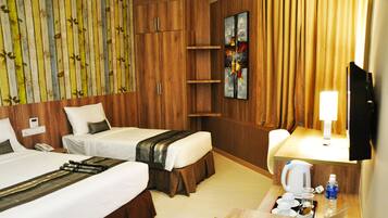 A hotel room with two beds, a wooden wardrobe, a painting, a television, and a small table with a kettle and water bottle.
