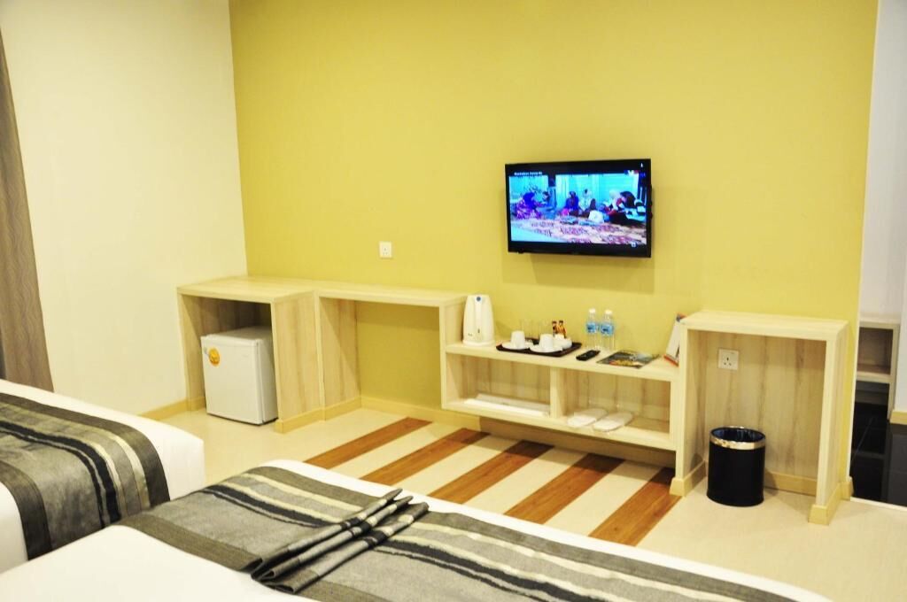 A hotel room with a television mounted on the wall, a small refrigerator, a bedside table with a lamp, a bedside chair, and a striped rug.