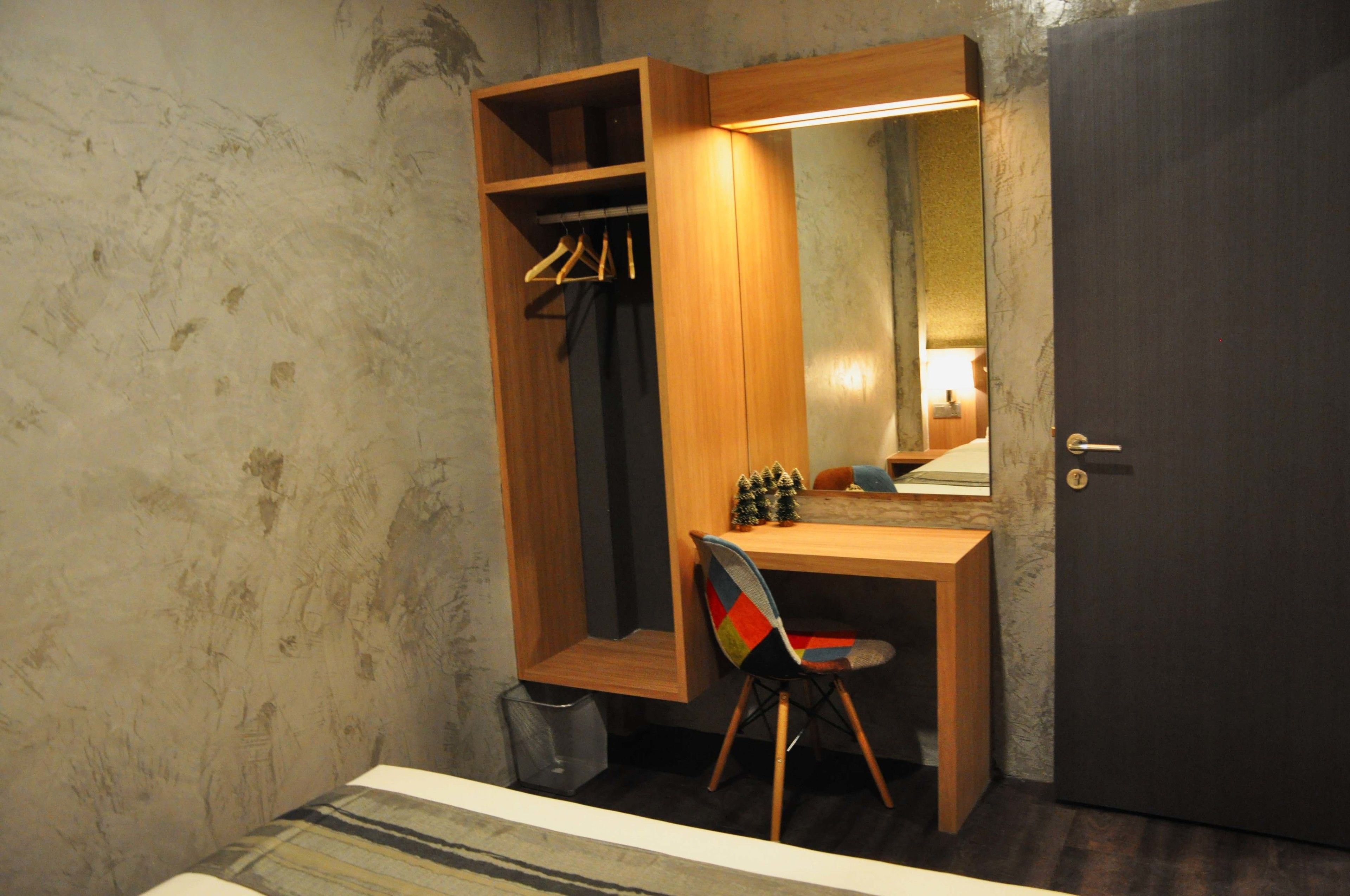 Una habitación pequeña y funcional con un escritorio de madera, una silla, un armario con ropa colgada y una puerta.