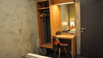 Una habitación pequeña y funcional con un escritorio de madera, una silla, un armario con ropa colgada y una puerta.