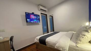 A hotel room with a bed, a desk, a chair, a TV mounted on the wall, and an air conditioning unit.