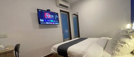 A hotel room with a bed, a desk, a chair, a TV mounted on the wall, and an air conditioning unit.