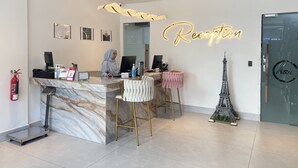 Uma área de recepção com balcão de mármore, dois bancos altos, um computador e um letreiro de neon com a palavra 'Reception'.