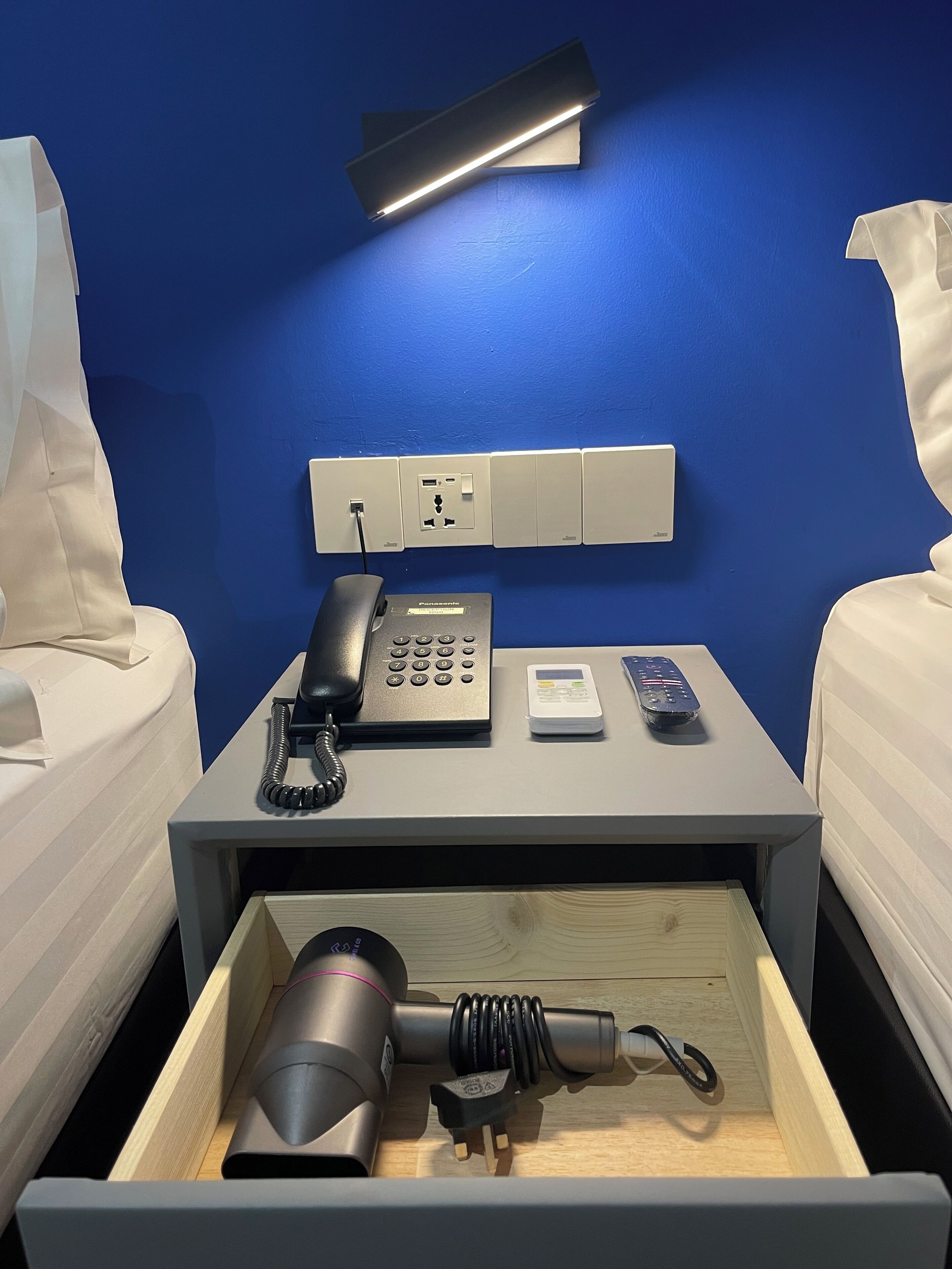 Habitación de hotel con cama, mesita de noche, teléfono, control remoto y un cajón con secador de pelo y otros objetos.