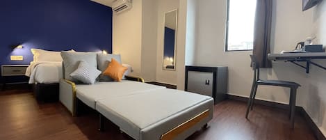 A hotel room with a bed, a desk, a chair, a small refrigerator, an air conditioning unit, and a window with curtains.