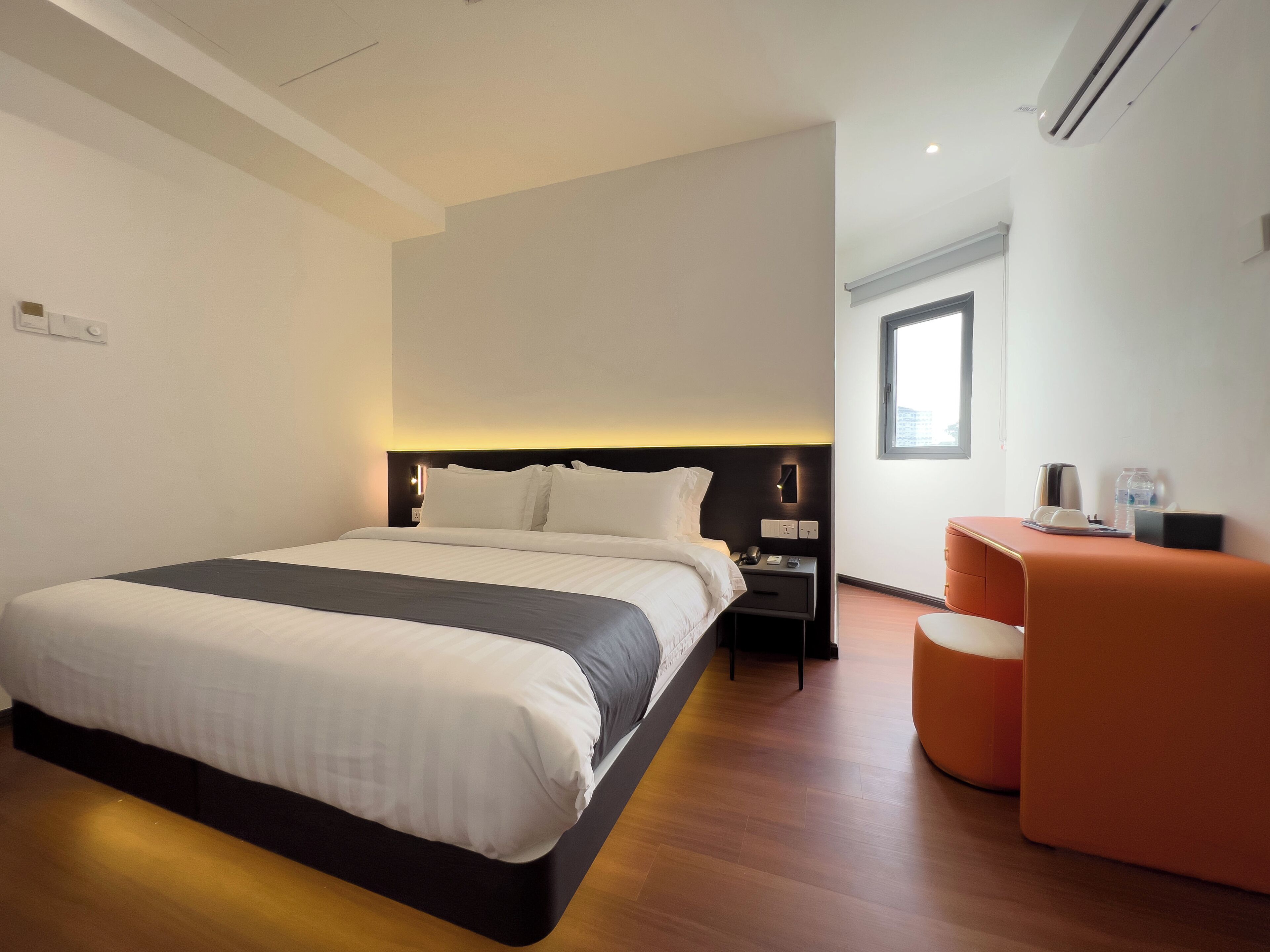 Una habitación de hotel moderna con una cama grande, una mesita de noche con lámpara, una zona de estar pequeña de color naranja y un ventanal que ofrece luz natural.