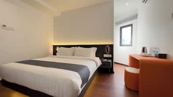 Una habitación de hotel moderna con una cama grande, una mesita de noche con lámpara, una zona de estar pequeña de color naranja y un ventanal que ofrece luz natural.