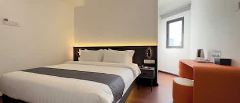 Una habitación de hotel moderna con una cama grande, una mesita de noche con lámpara, una zona de estar pequeña de color naranja y un ventanal que ofrece luz natural.